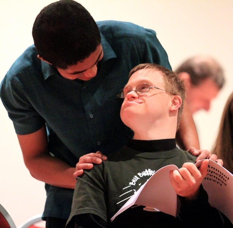 A man sits in a chair and looks up and behind himself. Another man stands over the chair and looks down at the man sitting. 