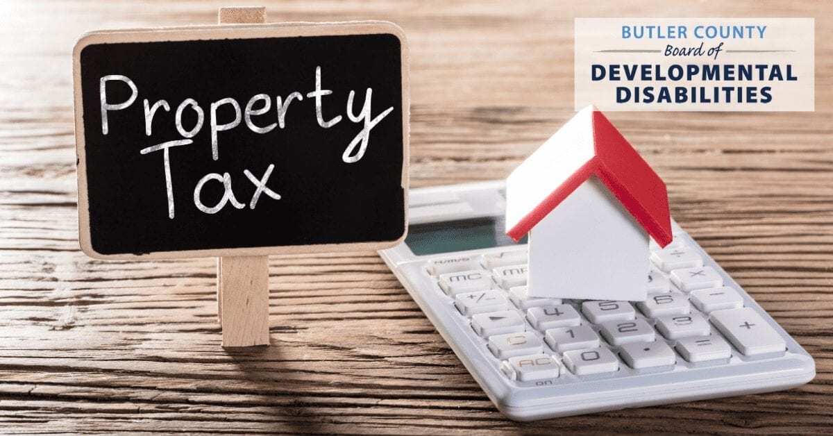 Set on a wooden table top: A small house sits on a calculator. A hand-made sign nearby reads "Property Tax." Superimposed on the image is the Butler County Board of Developmental Disabilities logo.