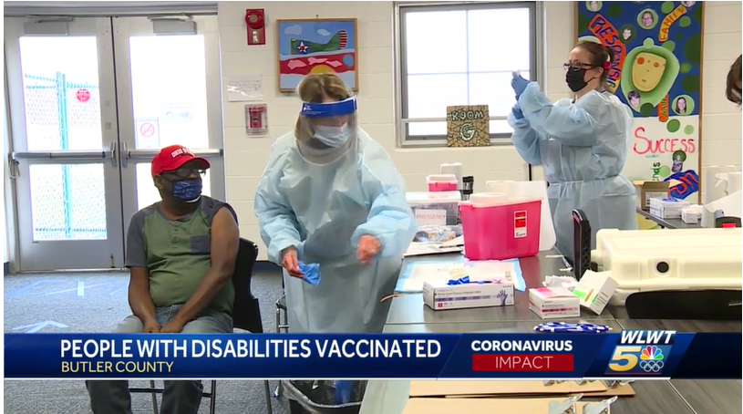 Two nurses work in full PPE while a man with a mask sits in a chair with his sleeve rolled up ready for a vaccine