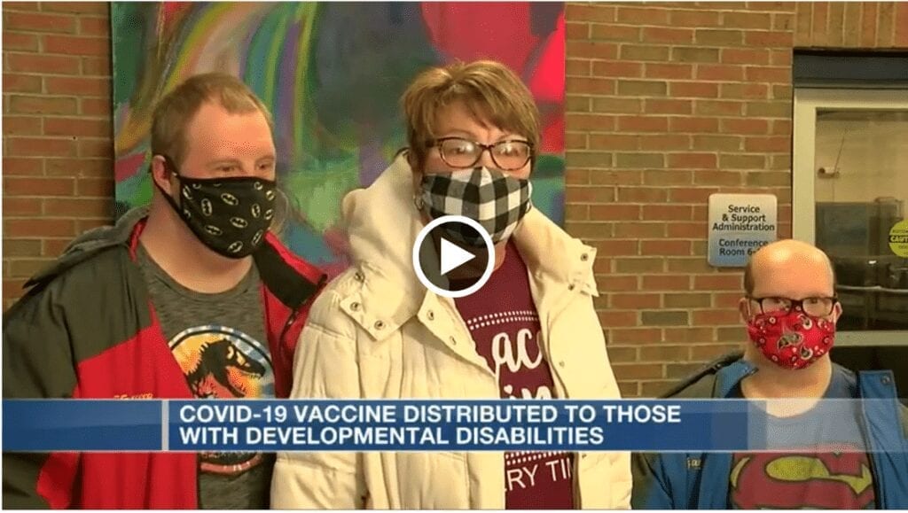 Tres personas de pie con máscaras. Un pie de foto dice: Vacuna COVID-19 distribuida a personas con discapacidades de desarrollo.  