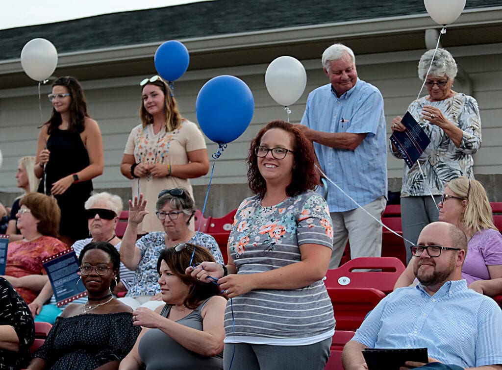 A woman stands holding a blue balloon. She is surrounded by other seated people.