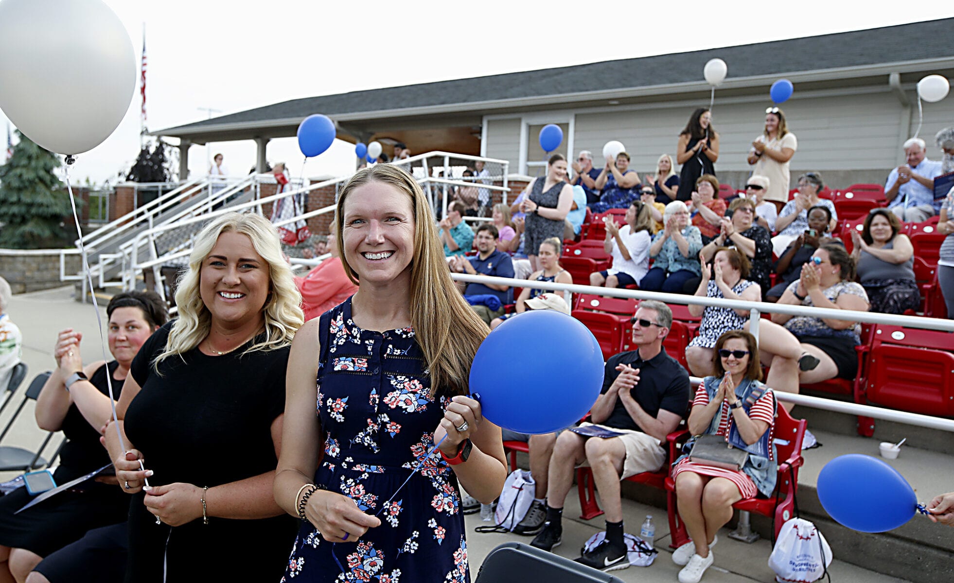 Two women, both holding balloons, smile at the camera. They stand in front of many seated guests at a ball park.