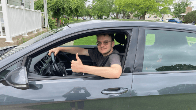 A young man sits in the driver's seat of a car with the window down. He is smiling at the camera and giving a thumbs up.