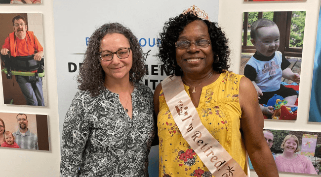 Two women stand side by side and smile at the camera. The woman on the right wears a tiara and a sash that reads "I am retired!"