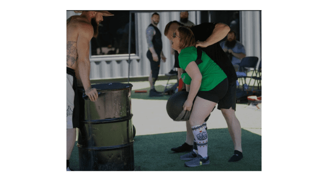 A woman strains to lift a heavily weighted ball as people in the background watch her performance. 