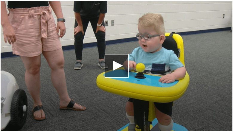 A toddler boy with glasses sits in a motorized chair. A play button is superimposed over the image.