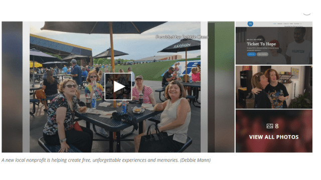 Women sit around a picnic table and smile at the camera. A play button is superimposed over the image indicating the image is a link to a video.