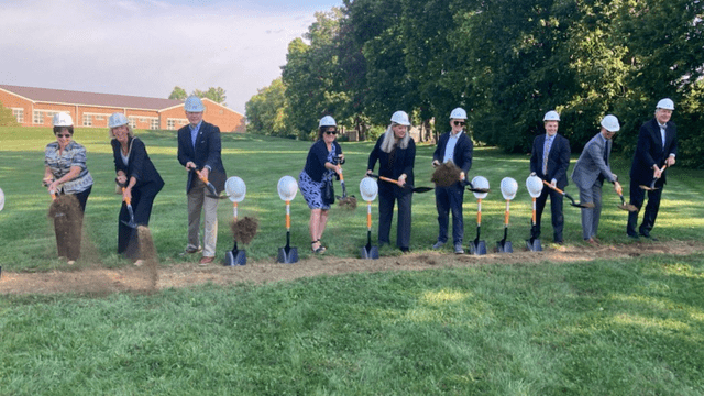 Nine professionally dressed people in hard hats sling dirt with ceremonial shovels.