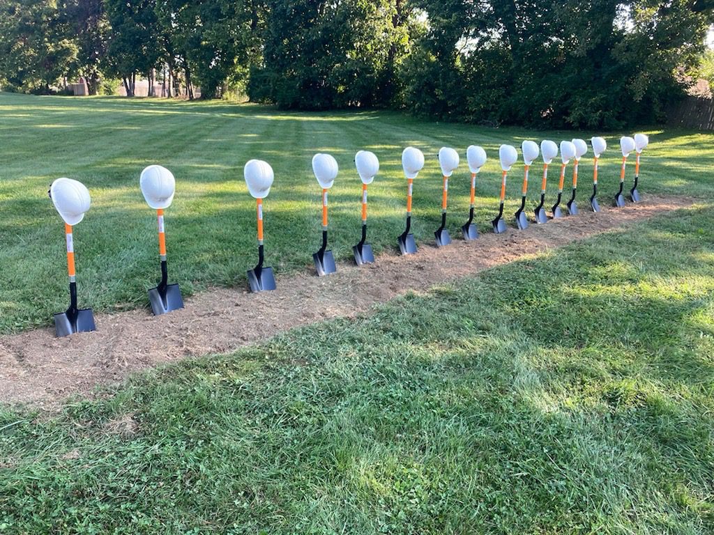 A row of shovels are stuck into freshly tilled dirt. A white hardhat rests upon hte the handle of each shovel.