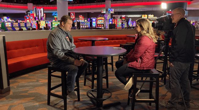 a man and woman sitting at a table across from each other indoors in front of a man with a camera filming them