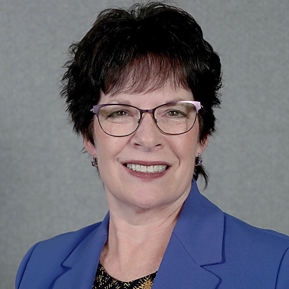 A headshot of woman with dark hair, glasses, and a blue suit jacket