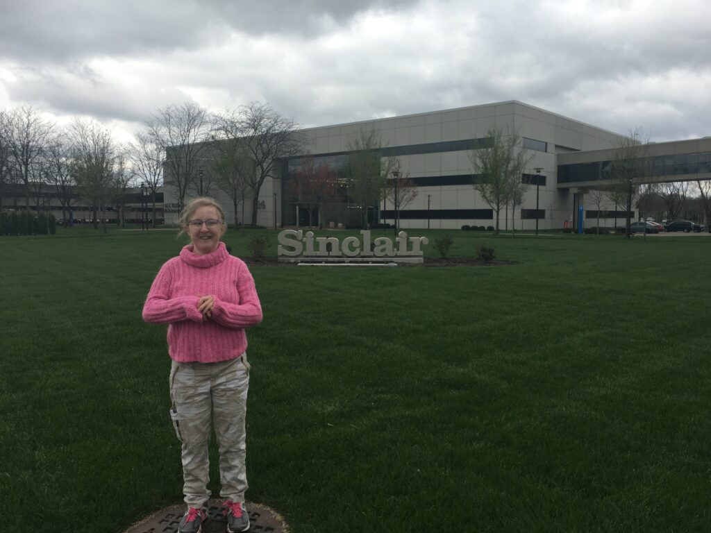 A woman outdoors in the grass with Sinclair Community College in the background.