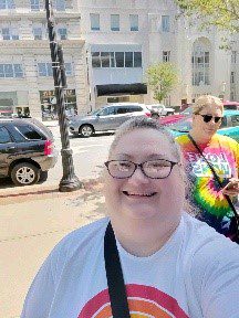 A woman wears a white shirt with a rainbow on it. She stands on a sidewalk and smiles at the camera. Behind her, someone wears a rainbow colored tie-die t-shirt. 