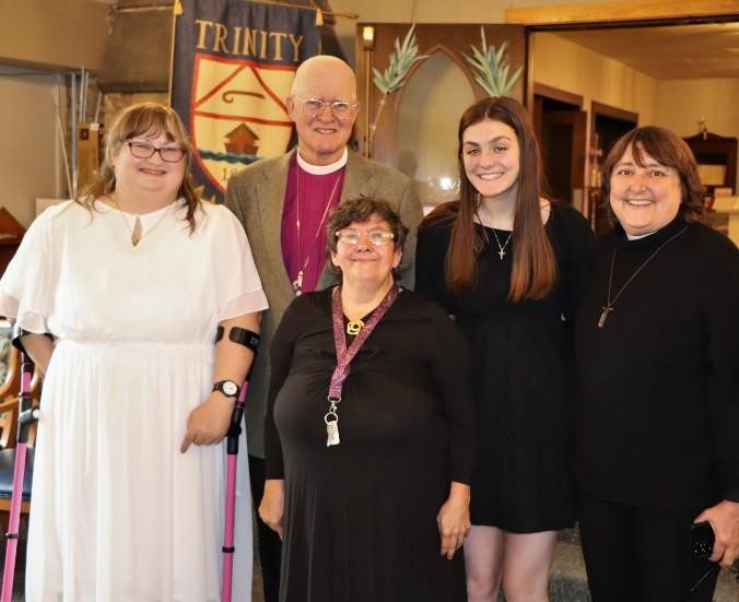 A group of four women and one man stand close together and smile at the camera. All are dressed in "church clothes" and appear to be standing in a church foyer. Three of them wear crucifixes around their necks. One woman uses crutches to help her stand. 