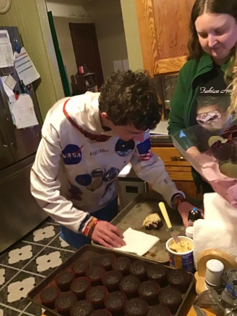 a man and a woman indoors standing at a table with baking supplies and cupcakes.