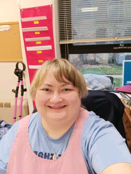 A woman sits at a desk and takes a joyful selfie. Behind her, files are organized on a hanging folder and pink crutches lean against the wall. 