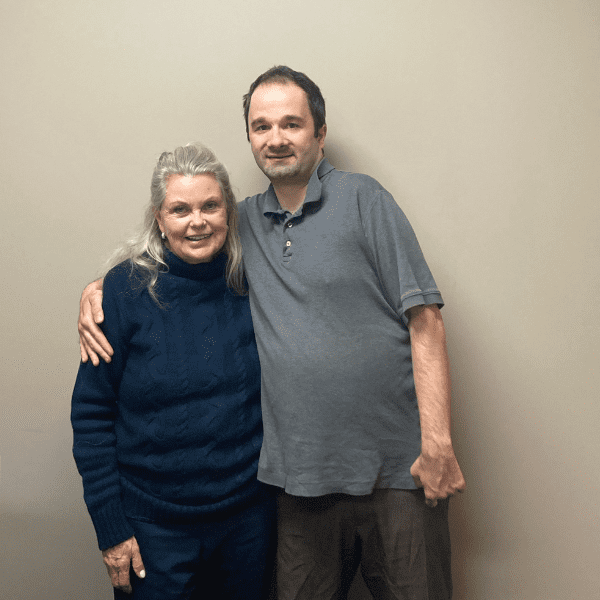 A woman and a man indoors against a beige wall standing side by side smiling