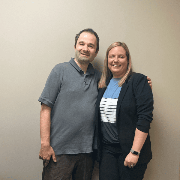 A man and a woman standing indoors against a beige wall side by side smiling