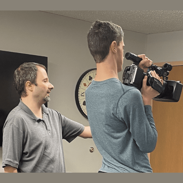 1 man teaching another man indoors in a classroom how to use a camera