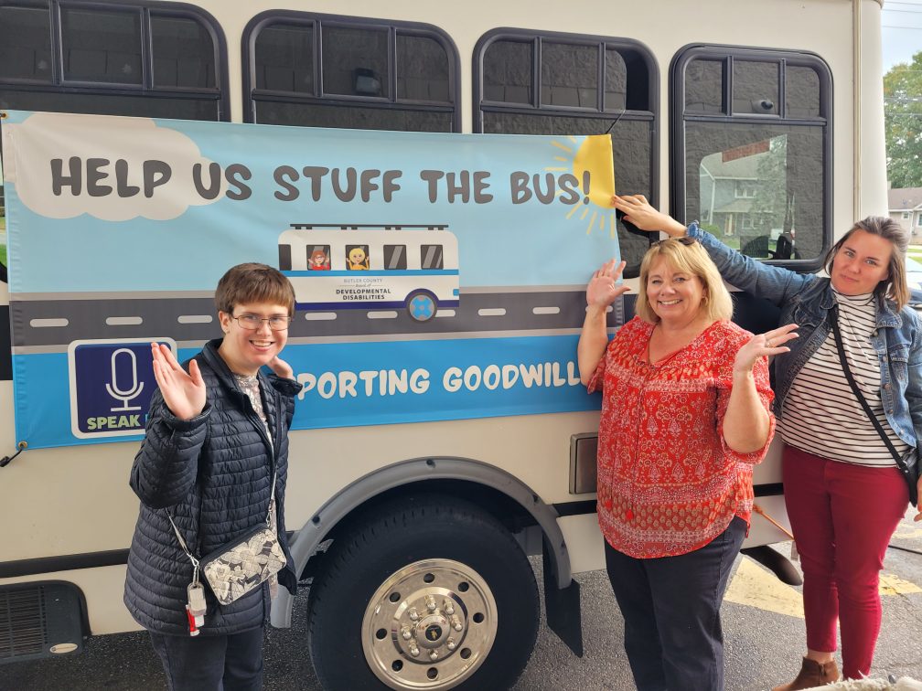 3 women standing outdoors in front of a bus that says, "Help Us Stuff the Bus, SpeakUP supporting Goodwill."