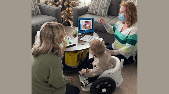 2 women and a baby in a mobility device indoors sitting at table using a tablet
