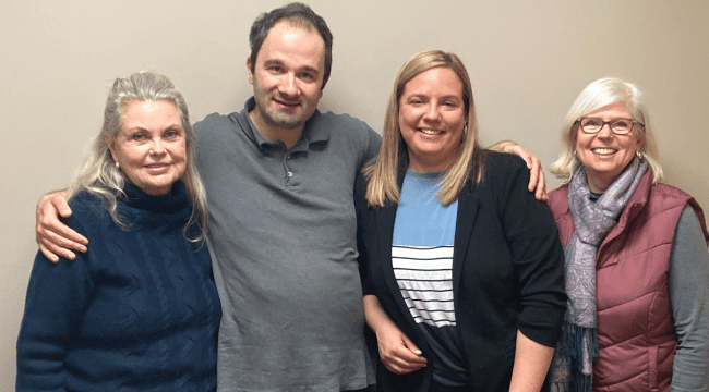 3 women and 1 man standing indoors together smiling for a photo