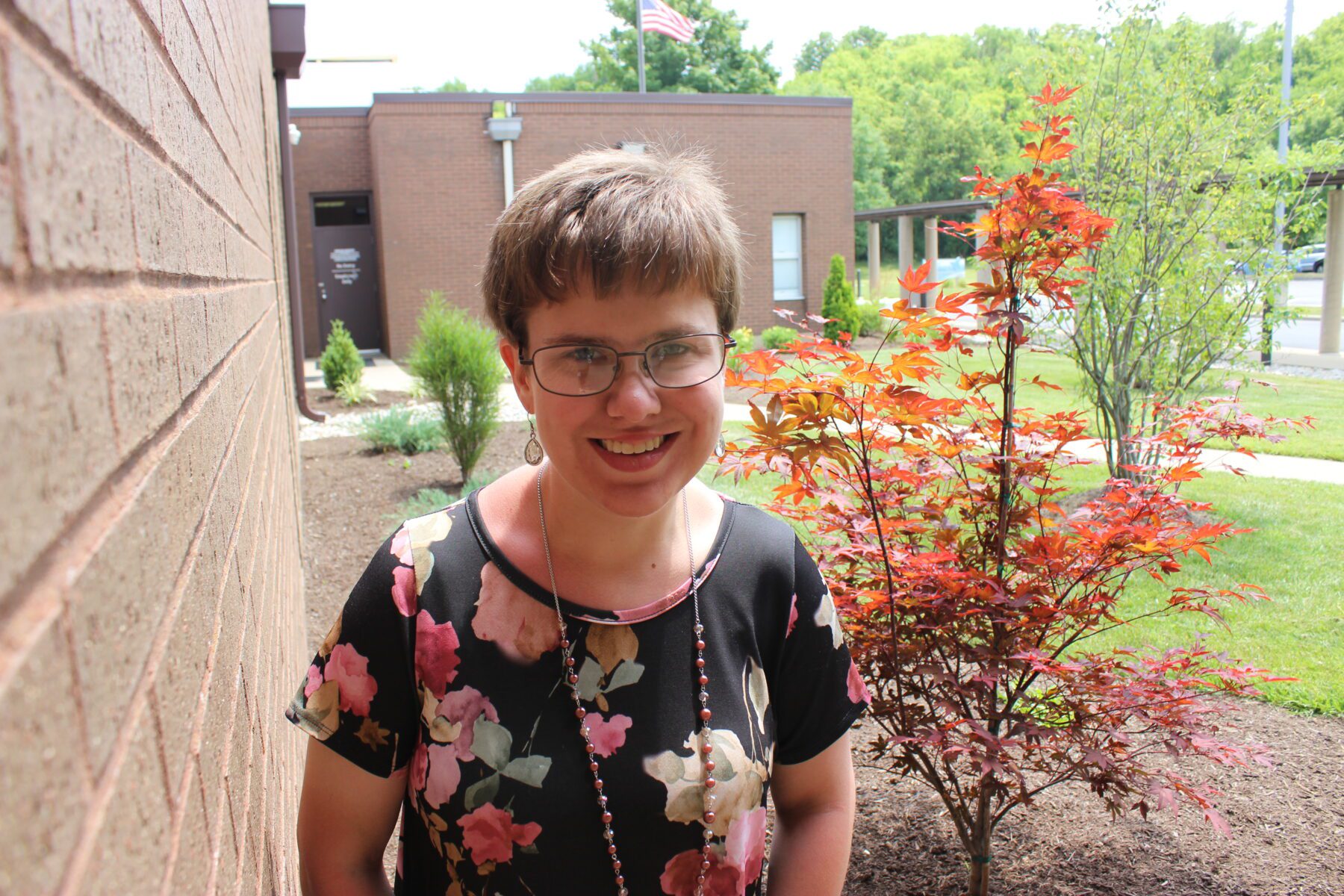 A young woman smiles brightly. She wears a floral shirt and stands with a brick wall on her left and landscaping on her right. Sun and shadows streak her smiling face. The image evokes a sense of brightness.