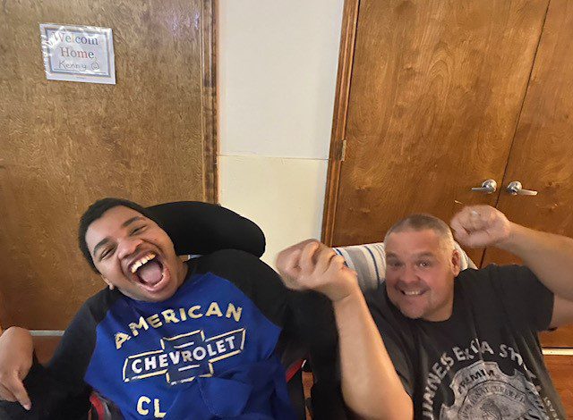 2 men indoors sitting in front of a door smiling together