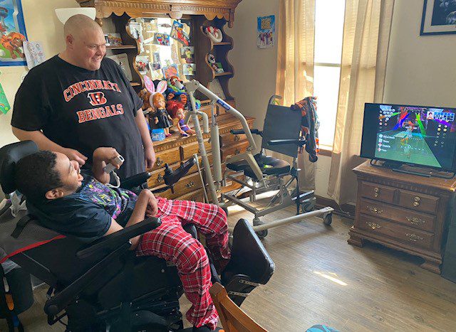 1 man standing next to a man in a wheelchair indoors watching television