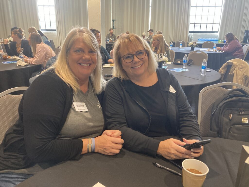 2 women indoors sitting at a table side by side