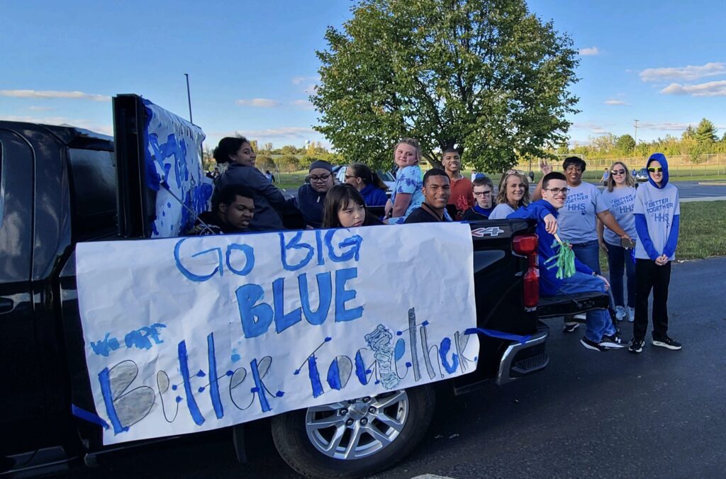 a group of students outdoors in parade sitting in a truck with a sign that says, "Go Big Blue, Better Together."
