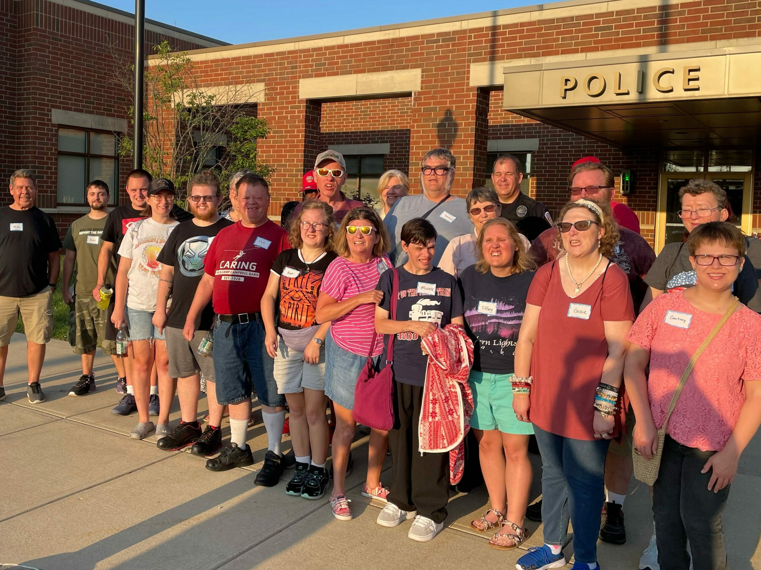 A large group of people stand outside and smile for the camera. They stand in front of a building with the word Police above the door. 