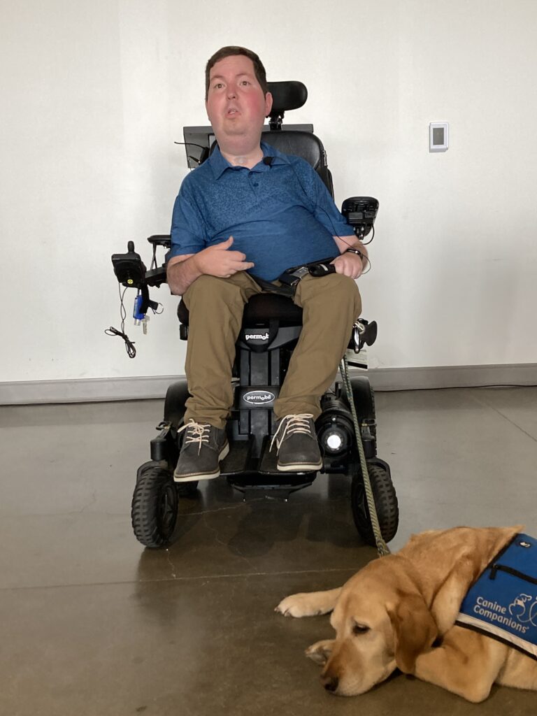 A man sits in a motorized wheelchair. A service dog lays at his feet. 