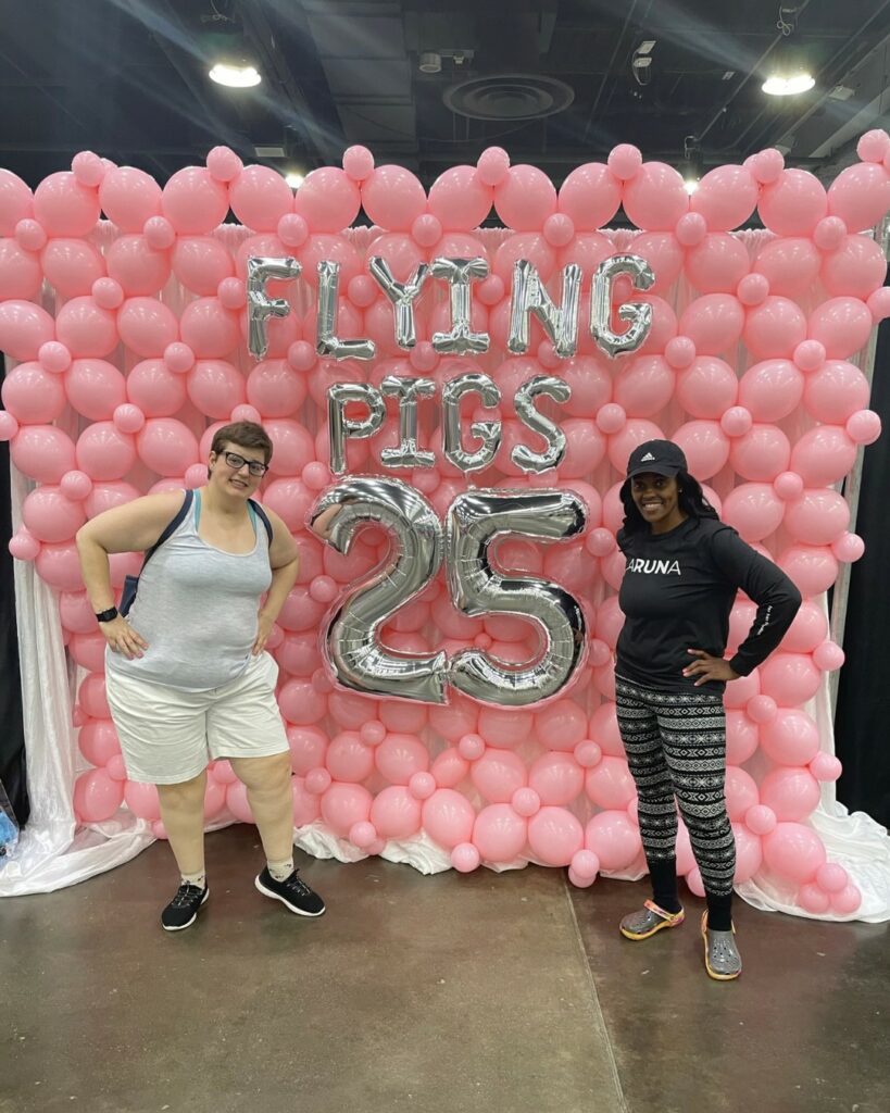 2 women indoors standing in front of pink balloons and silver balloons that spell, "Flying Pigs 25."