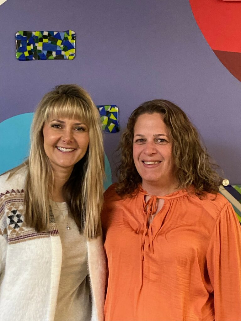 2 women indoors in front of a display of glass art standing side by side smiling