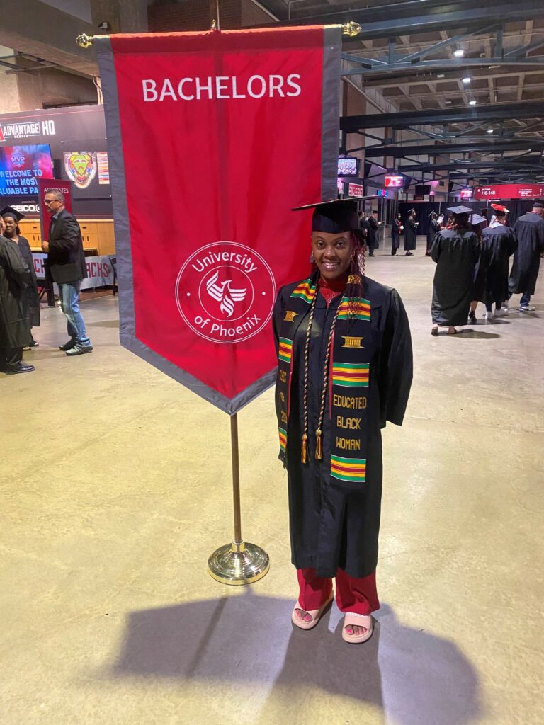 a women in a graduate uniform indoors in front of a banner that says Bachelors University of Phoenix