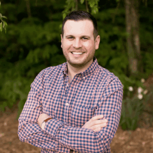 A man stands on a wooded path. He has his arms crossed and he is smiling at the camera. He wears a red and blue plaid button down shirt. 