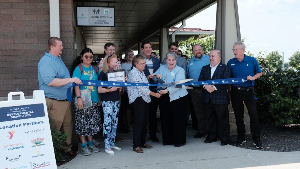 Many people stand behind a ribbon. Two women in the middle hold a large pair of scissors and begin to cut the ribbon. Above the group hangs a sign that says Family restroom Universal Changing tables.