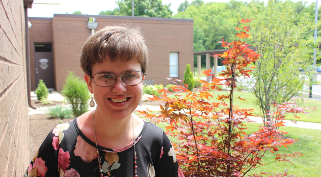 A young woman stands outside on a sunny day. She wears a black floral shirt and glasses. She is smiling directly at the camera.
